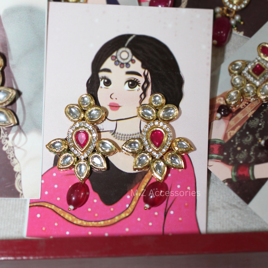 Gold-toned traditional Indian earrings with red heart-shaped stones and clear rhinestone floral borders, displayed on a white card featuring an illustrated woman in a pink outfit. The card is surrounded by other jewelry pieces.