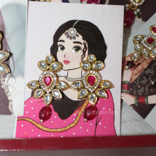 Gold-toned traditional Indian earrings with red heart-shaped stones and clear rhinestone floral borders, displayed on a white card featuring an illustrated woman in a pink outfit. The card is surrounded by other jewelry pieces.